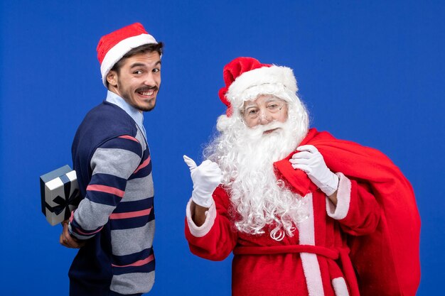 Vue de face du père noël avec un jeune homme sac de transport plein de cadeaux sur les vacances bleues présente la couleur de noël