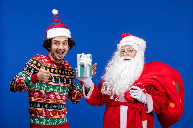 Vue de face du père noël avec un jeune homme tenant un cadeau de noël sur un mur bleu