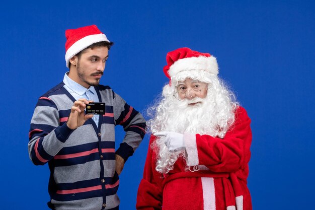 Vue de face du père noël avec un jeune homme tenant une carte bancaire sur le mur bleu