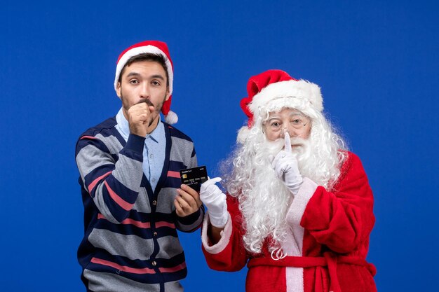 Vue de face du père noël avec un jeune homme tenant une carte bancaire sur le mur bleu