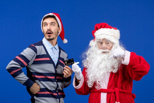 Vue de face du père noël avec un jeune homme tenant une carte bancaire sur le mur bleu