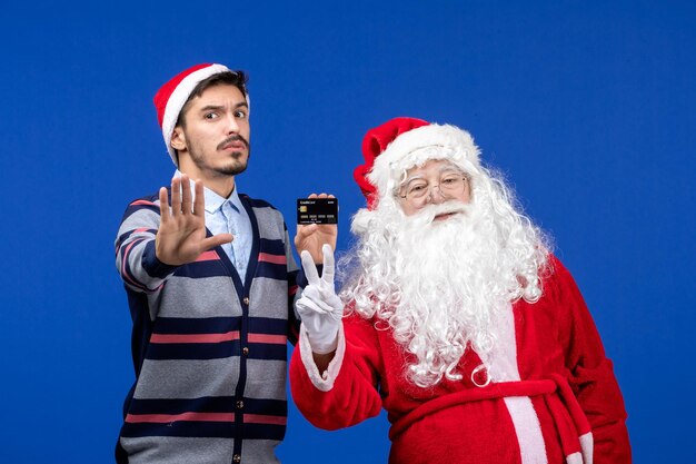 Vue de face du père noël avec un jeune homme tenant une carte bancaire sur le mur bleu