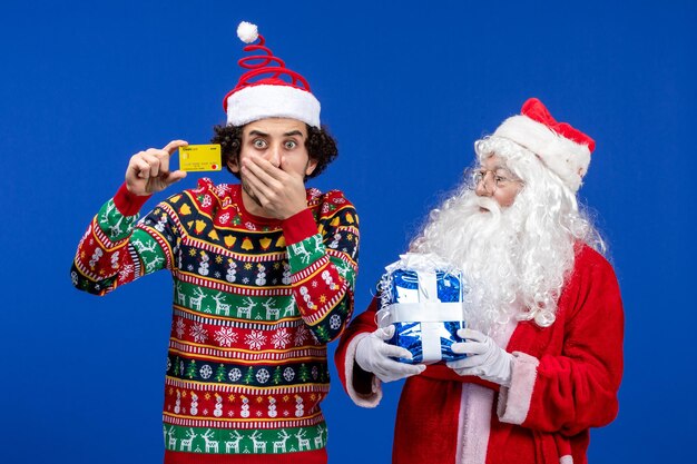 Vue de face du père noël avec un jeune homme tenant une carte bancaire et présent sur le mur bleu