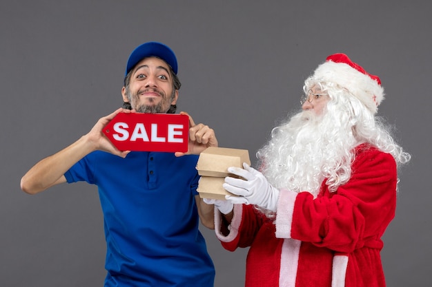 Photo gratuite vue de face du père noël avec messager masculin tenant la bannière de vente et les emballages alimentaires sur le mur gris