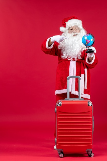 Vue de face du père Noël avec sac tenant petit globe et préparation pour le voyage sur le mur rouge