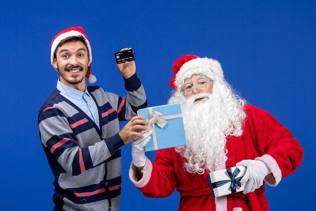 Vue de face du père noël tenant des cadeaux et jeune homme tenant une carte bancaire sur la couleur des vacances du nouvel an bleu noël