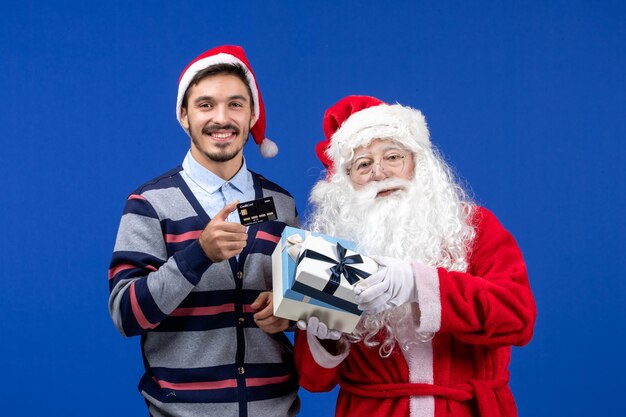 Vue de face du père noël tenant des cadeaux et jeune homme tenant une carte bancaire pendant les vacances de noël bleu