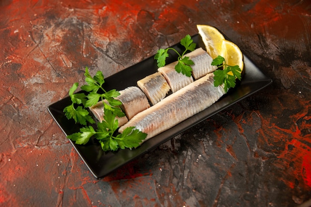 Vue de face du poisson frais tranché avec des légumes verts et des morceaux de citron à l'intérieur d'une casserole noire sur de la viande de collation sombre