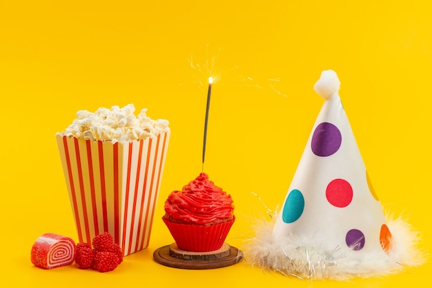 Une vue de face du pop-corn et du gâteau avec chapeau d'anniversaire et marmelades sur bureau jaune