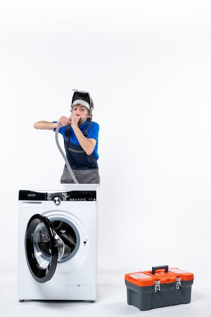 Vue de face du réparateur aux yeux écarquillés en uniforme debout derrière la machine à laver soufflant le tuyau sur un mur isolé blanc