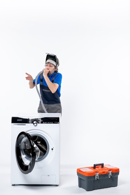 Vue de face du réparateur confus en uniforme debout derrière la machine à laver soufflant le tuyau sur le mur blanc