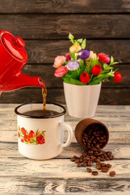 Vue de face du thé chaud coulant de la bouilloire rouge avec des graines de café brun et des fleurs sur le bureau en bois