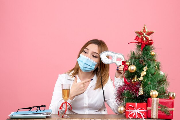 Vue de face du travailleur féminin en masque assis derrière sa table avec des cadeaux de Noël sur rose