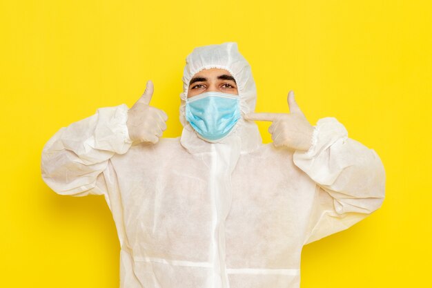 Vue de face du travailleur scientifique masculin en combinaison blanche de protection spéciale avec masque sur le mur jaune clair