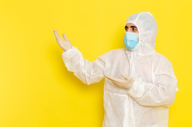 Vue de face du travailleur scientifique masculin en combinaison blanche de protection spéciale avec masque sur le mur jaune clair