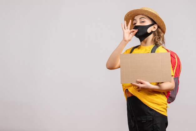 Vue de face du voyageur féminin avec sac à dos tenant le carton faisant signe savoureux sur mur gris