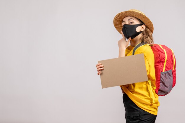 Vue de face du voyageur féminin avec sac à dos tenant le carton mettant la main sur son menton sur un mur gris