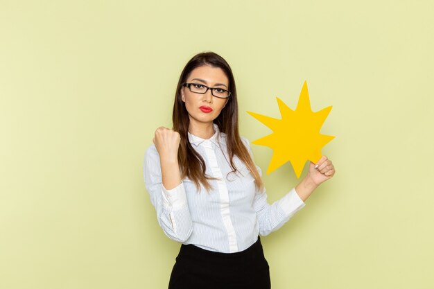 Vue de face de l'employée de bureau en chemise blanche et jupe noire tenant une pancarte jaune sur un mur vert clair