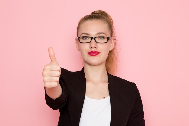 Vue de face de l'employée de bureau en veste noire stricte montrant comme signe sur mur rose clair