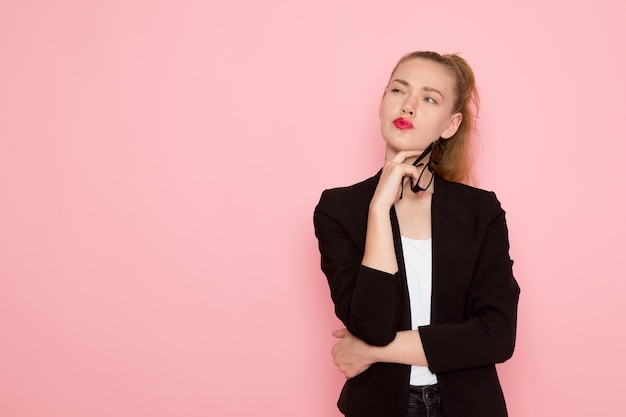 Vue de face de l'employée de bureau en veste noire stricte posant avec expression de pensée sur le mur rose