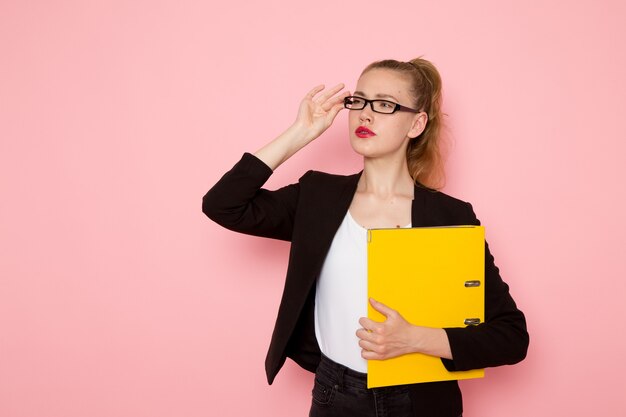 Vue de face de l'employée de bureau en veste noire stricte tenant des fichiers jaunes sur un mur rose clair