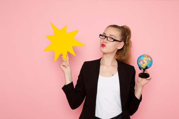 Vue De Face De L'employée De Bureau En Veste Noire Stricte Tenant Un Grand Panneau Jaune Avec Globe Sur Mur Rose