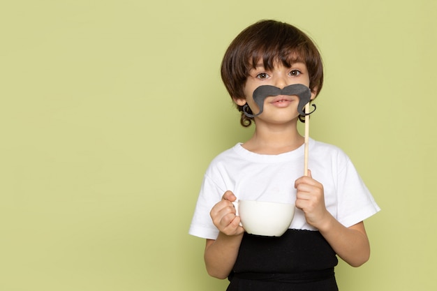 Photo gratuite une vue de face enfant garçon en t-shirt blanc tenant la moustache et une tasse de café sur l'espace de couleur pierre