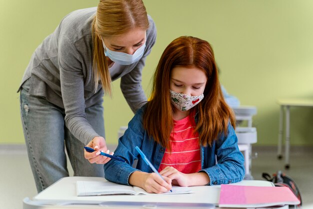 Vue de face de l'enseignante avec masque médical aidant la petite fille en classe