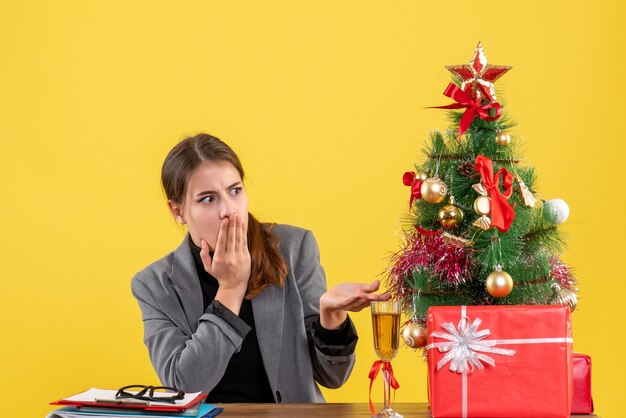 Vue de face a erré fille assise à la table de Noël arbre et cadeaux cocktail