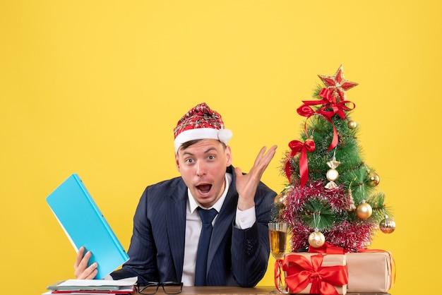 Vue de face étonné homme d'affaires assis à la table près de l'arbre de Noël et présente sur fond jaune