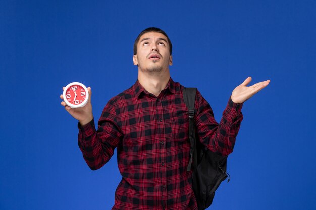 Vue de face de l'étudiant en chemise à carreaux rouge avec sac à dos tenant des horloges sur le mur bleu