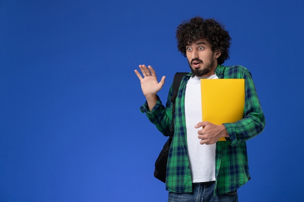 Vue de face de l'étudiant en chemise à carreaux vert portant un sac à dos noir et tenant des fichiers sur le mur bleu
