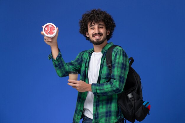 Vue de face de l'étudiant en chemise à carreaux vert portant un sac à dos noir et tenant des horloges et du café sur le mur bleu