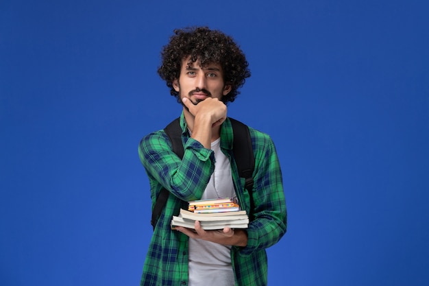 Photo gratuite vue de face de l'étudiant en chemise à carreaux vert avec sac à dos noir tenant des cahiers et des fichiers sur le mur bleu