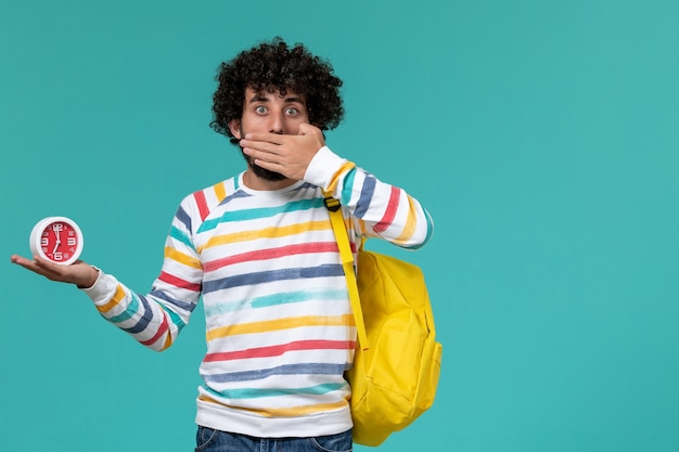 Photo gratuite vue de face de l'étudiant en chemise rayée portant un sac à dos jaune tenant des horloges couvrant sa bouche sur le mur bleu