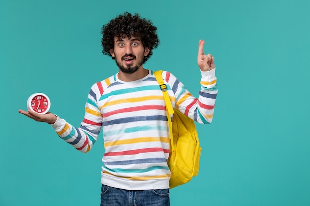 Vue de face de l'étudiant en chemise rayée portant un sac à dos jaune tenant des horloges croisant ses doigts sur le mur bleu