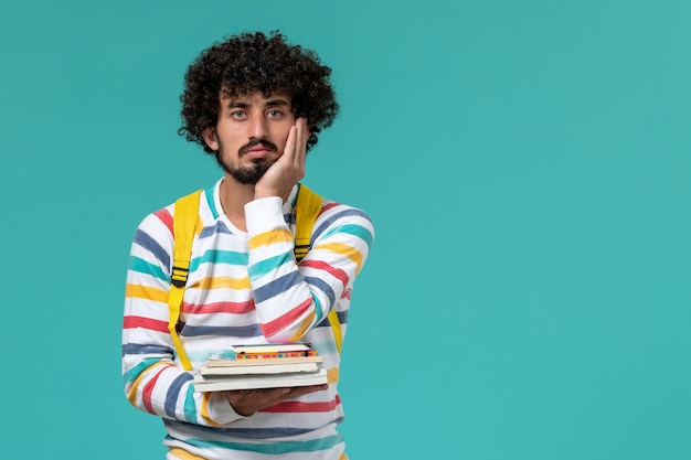 Vue de face de l'étudiant en chemise rayée portant un sac à dos jaune tenant des livres pensant sur le mur bleu