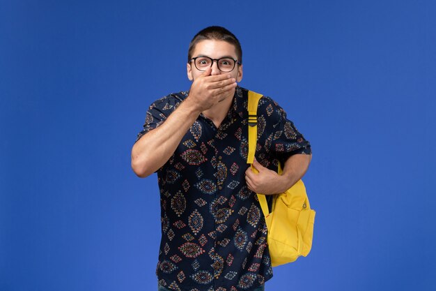 Vue de face de l'étudiant en chemise sombre portant un sac à dos jaune fermant la bouche sur un mur bleu clair