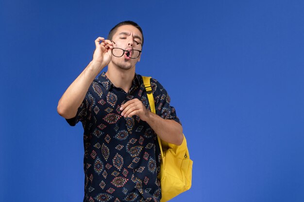 Vue de face de l'étudiant en chemise sombre portant un sac à dos jaune nettoyant ses lunettes de soleil sur un mur bleu clair