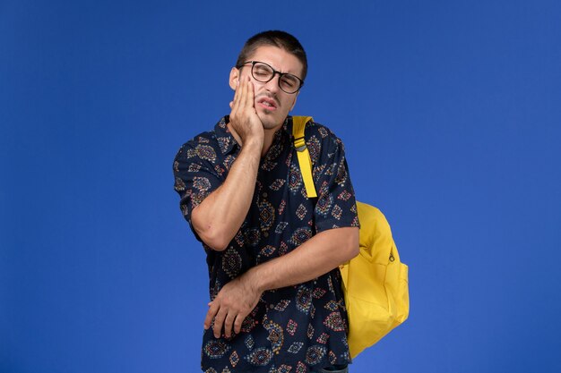 Vue de face de l'étudiant en chemise sombre portant un sac à dos jaune souffrant de maux de dents sur un mur bleu clair