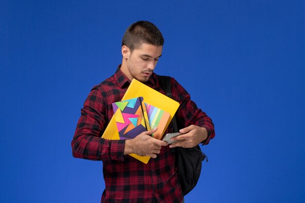 Vue de face de l'étudiant masculin en chemise à carreaux rouge avec sac à dos tenant un cahier et des fichiers à l'aide de son téléphone sur le mur bleu