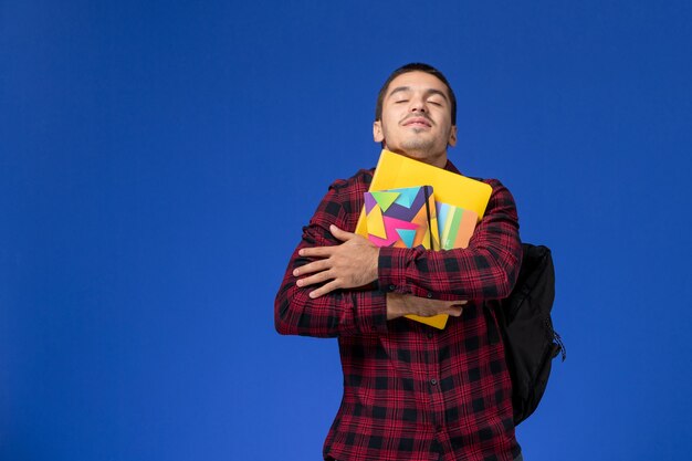 Vue de face de l'étudiant masculin en chemise à carreaux rouge avec sac à dos tenant un cahier et des fichiers sur le mur bleu clair