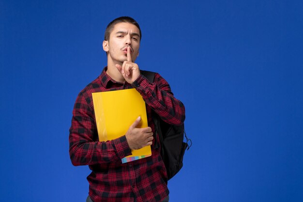 Vue de face de l'étudiant masculin en chemise à carreaux rouge avec sac à dos tenant des fichiers jaunes sur le mur bleu