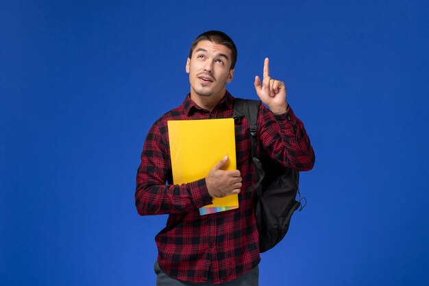 Vue de face de l'étudiant masculin en chemise à carreaux rouge avec sac à dos tenant des fichiers jaunes sur le mur bleu