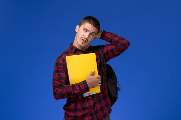 Vue de face de l'étudiant masculin en chemise à carreaux rouge avec sac à dos tenant des fichiers jaunes pensant sur le mur bleu