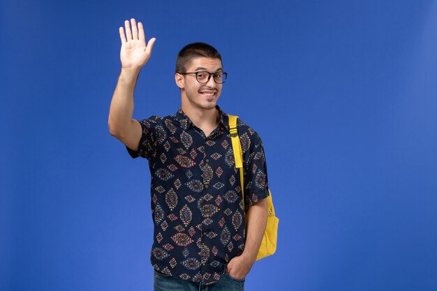 Vue de face de l'étudiant masculin en chemise de coton foncé portant un sac à dos jaune en agitant sa main sur le mur bleu
