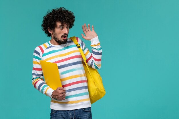 Vue de face de l'étudiant masculin en chemise rayée portant sac à dos jaune tenant des fichiers sur le mur bleu