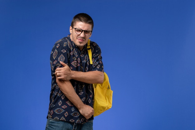 Vue de face de l'étudiant masculin en chemise sombre portant un sac à dos jaune sur le mur bleu