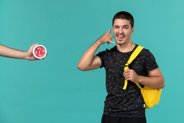 Vue de face de l'étudiant masculin en t-shirt foncé sac à dos jaune posant sur le mur bleu clair
