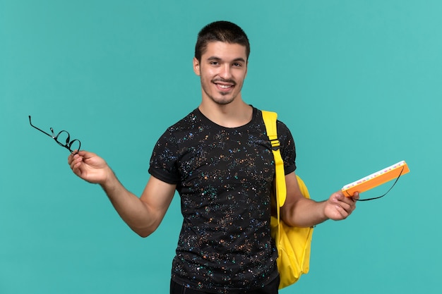 Vue de face de l'étudiant masculin en t-shirt foncé sac à dos jaune tenant un cahier sur le mur bleu